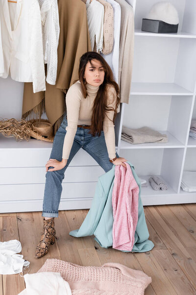 Pensive woman holding clothes while sitting on shelf in wardrobe 