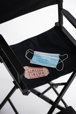 Tıbbi maskeler ve sinema biletleri beyaz sinema konsepti üzerine film yapımcısı sandalyesinde yüksek açılı görünüyor.