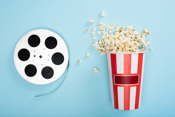 top view of film bobbin near bucket of popcorn on blue, cinema concept