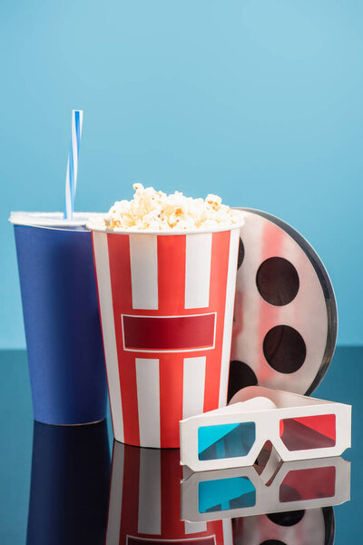 bucket with popcorn, cup with soda, film bobbin and 3d cinema glasses on glossy surface isolated on blue