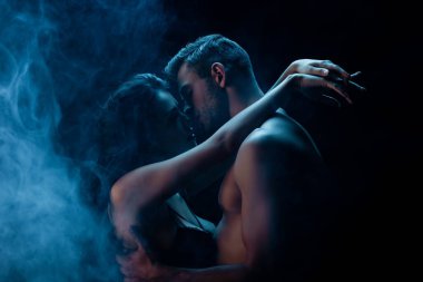 Siyah arka planda sütyenli kız arkadaşını dumanla öpen seksi bir adam. 