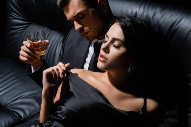 Kanepede, şehvetli bir kadının yanında elinde viski bardağı tutan bir adam. 