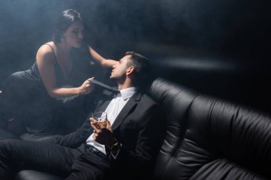 Seksi kadın takım elbiseli erkek arkadaşının kravatını tutuyor siyah arka planda bir bardak viski tutuyor dumanlı. 
