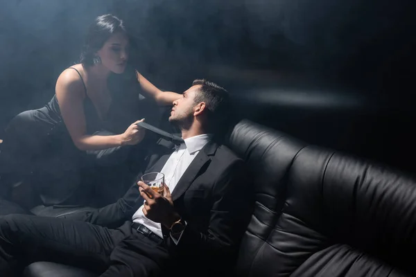 Sexy woman holding tie of boyfriend in suit holding glass of whiskey on black background with smoke