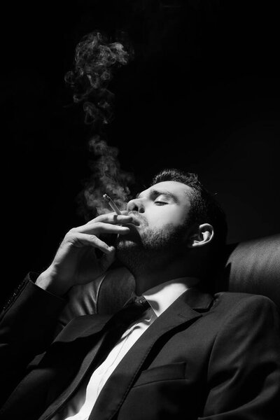 Monochrome shot of young man in suit smoking cigarette isolated on black 