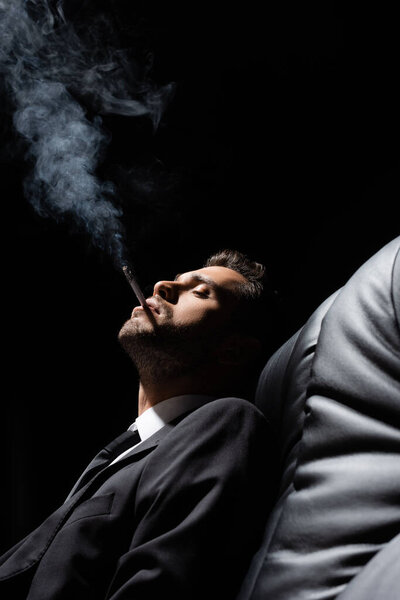 Молодой человек в костюме курит сигарету на диване на размытом переднем плане, изолированном на черном 