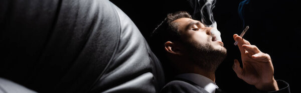 Человек в костюме курит сигарету на диване изолирован на черном, баннер 
