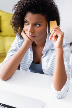 Pahalı Afro-Amerikalı kadın, ön planda bulanık dizüstü bilgisayarın yanında kredi kartı tutuyor. 