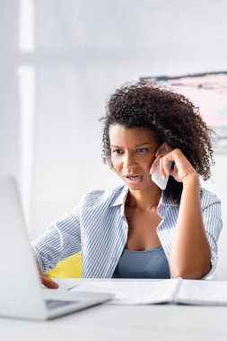 Bulanık ön planda dizüstü bilgisayar kullanan ve akıllı telefondan konuşan Afrikalı Amerikalı serbest yazar. 