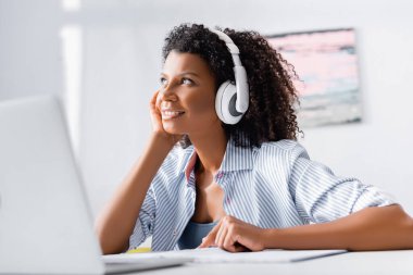 Afro-Amerikalı kadın kulaklıkla kulaklıkla ön planda bulanık bir şekilde müzik dinlerken gülümsüyor. 