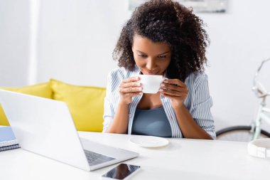 Önplanda, bilgisayarın ve akıllı telefonun yanında fincanı tutan Afrikalı Amerikalı kadın. 