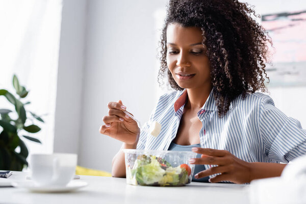 African american woman looking at salad near cup on blurred foreground 