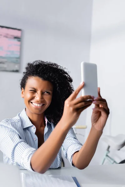 Positive african american woman winking while taking selfie near notebook
