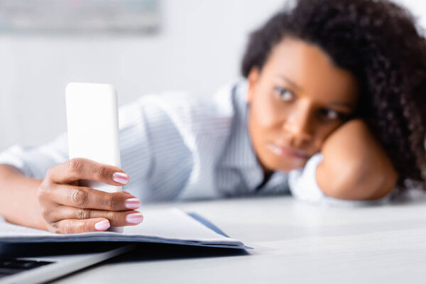 Смартфон в руке африканской американки на размытом фоне возле ноутбука и ноутбука 