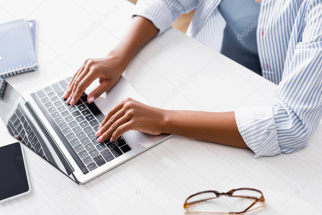 Cropped view of african american teleworker using laptop near eyeglasses and smartphone 
