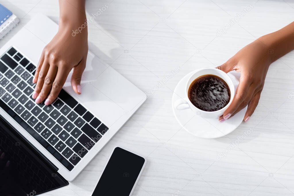 Cropped view of african american woman using laptop near smartphone with blank screen and holding cup of coffee 