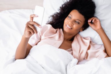 Yatakta bulanık arka planda gülümseyen Afrikalı Amerikalı bir kadının elinde akıllı telefonun yüksek açılı görüntüsü 