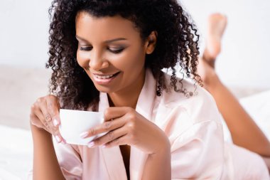 Positive african american woman looking at cup of coffee in bedroom  clipart