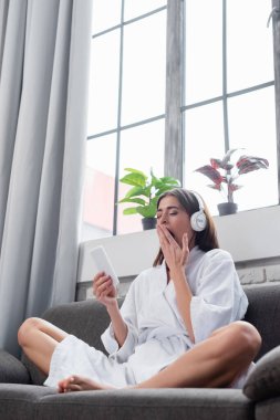 Uykulu, genç, yetişkin bir kadın bacak bacak üstüne atmış ve oturma odasındaki kanepede kulaklıkla müzik dinliyor.