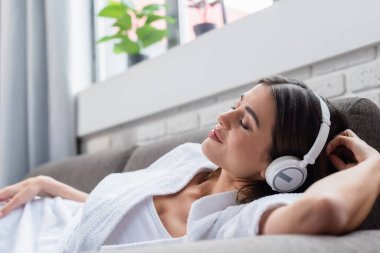 peaceful young adult woman with closed eyes resting in headphones on couch at home clipart