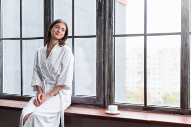 pretty young adult woman in bathrobe sitting on sill with cup of coffee near window at home clipart