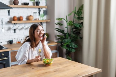 Bornozlu genç bir kadın salata yiyor ve modern mutfakta cep telefonuyla konuşuyor.
