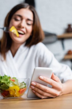 Modern mutfakta bornozlu, salata yiyen ve cep telefonu kullanan neşeli genç kadın ön plana odaklan.