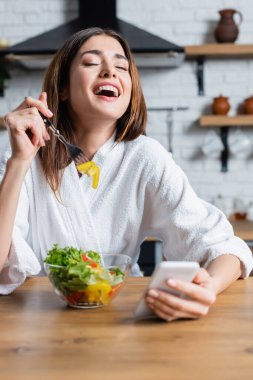 Bornozlu genç bir kadın sebze yiyor salata yiyor ve modern mutfakta cep telefonu kullanıyor.