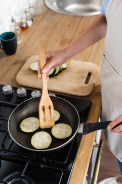 cropped view of young adult woman cooking slices of eggplant in frying pan in kitchen clipart