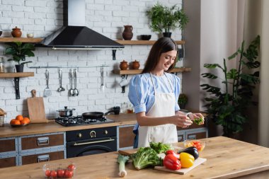 Önlüklü mutlu yetişkin kadın modern mutfakta sebze salatası hazırlıyor.