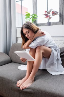 Kanepede oturan ve modern oturma odasındaki tablete bakan genç bir kadın.