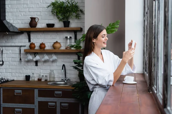 Glimlachende Jonge Volwassen Vrouw Badjas Messaging Mobiele Telefoon Buurt Venster — Stockfoto