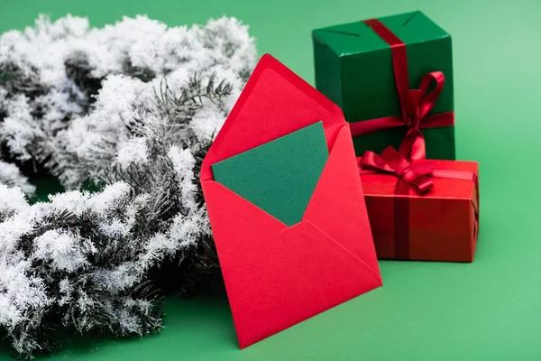 Envelope vermelho com cartão perto de caixas de presente e ramo de abeto com neve decorativa no verde — Fotografia de Stock