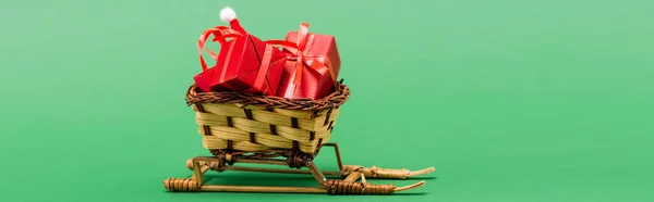 Encabezado del sitio web de cajas de regalo rojas y sombrero de santa en canasta de mimbre en trineo decorativo sobre fondo verde - foto de stock
