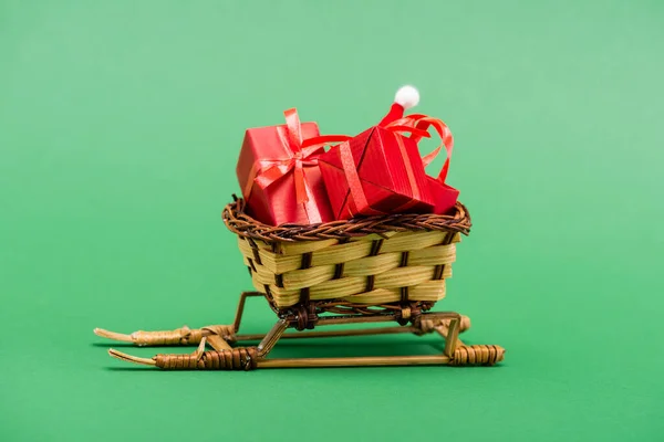 Rote Geschenkboxen und Weihnachtsmütze im Weidenkorb auf dekorativem Schlitten auf grünem Hintergrund — Stockfoto