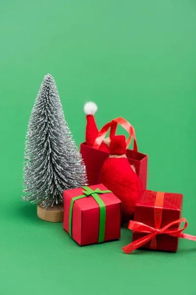 Decorative silver christmas tree near gift boxes, sack and shopping bag with santa hat on green — Stock Photo