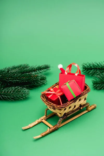 Geschenkboxen und Einkaufstasche mit Weihnachtsmann im Weidenkorb auf dekorativem Schlitten in der Nähe von Tannenzweigen auf Grün — Stockfoto