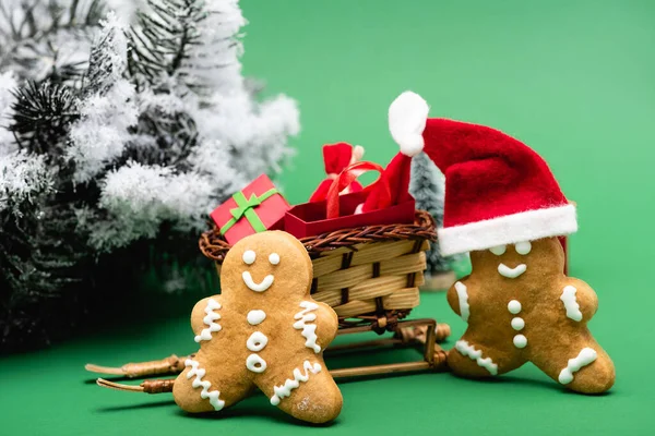 Lebkuchenmänner in der Nähe von Schlitten mit Einkaufstasche und Geschenkbox und Tannenzweig mit dekorativem Schnee auf grünem Hintergrund — Stockfoto