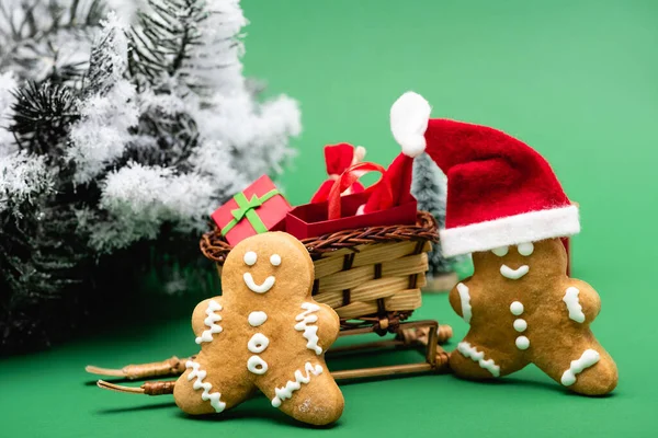 Hommes de pain d'épice près traîneau avec sac à provisions et boîte cadeau, et branche de pin avec neige décorative sur fond vert — Photo de stock