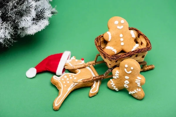 Lebkuchenmänner und Hirsche mit Weihnachtsmütze neben dekorativem Schlitten und Tannenzweig mit Schnee auf grünem Grund — Stockfoto