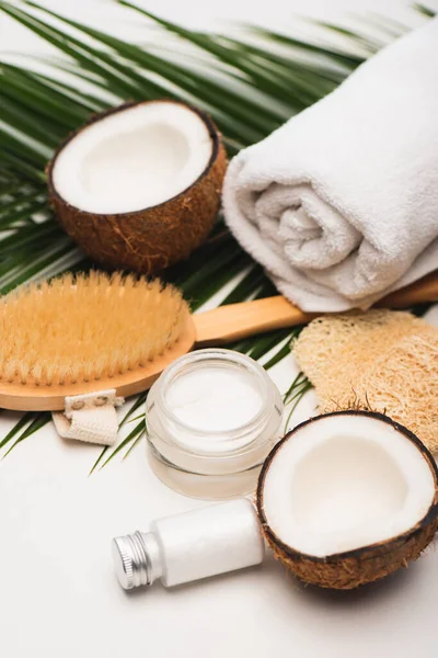 Moitiés de noix de coco, crème cosmétique maison et lotion près de serviette, brosse de massage et feuilles de palmier sur blanc — Photo de stock