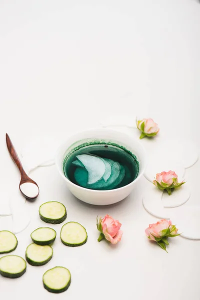 Bowl with tonic and cotton pads, cucumber slices, tea roses, and spoon with cosmetic cream on white — Stock Photo