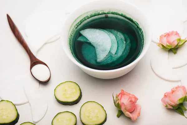 Bowl with cleansing lotion and cotton pads near sliced cucumber, tea roses, and wooden spoon with cosmetic cream on white — Stock Photo