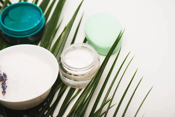 Recipientes com creme cosmético e esfoliação, sabão e tigela com galho de lavanda perto de folhas de palmeira em branco — Fotografia de Stock