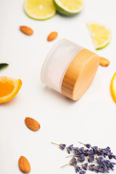 Container of homemade cosmetic cream near sliced orange, lime and lemon, almonds and lavender twigs on white blurred background — Stock Photo