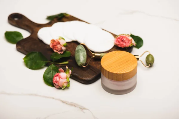 Container with cosmetic cream near cotton pads, tea roses, and jade roller on white marble background — Stock Photo