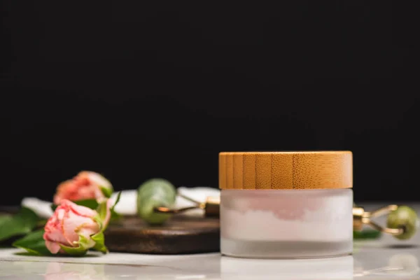 Container with homemade cosmetic cream near jade roller and tea roses on chopping board on blurred background isolated on black — Stock Photo