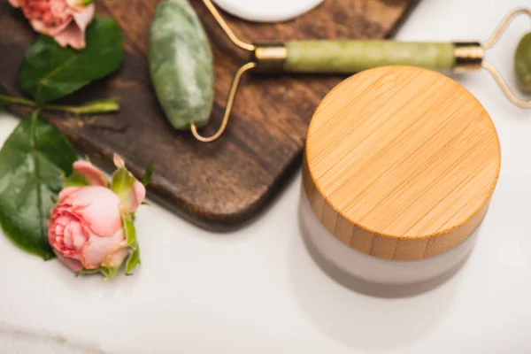 Container of cosmetic cream near chopping board with jade roller, and tea roses on blurred white background — Stock Photo