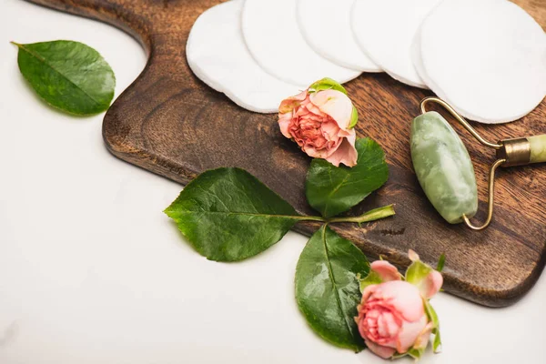 Tabla de cortar de madera con rosas de té, almohadillas de algodón y rodillo de jade sobre fondo blanco - foto de stock