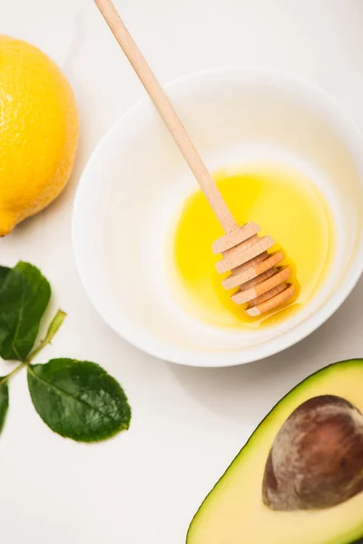 Vue du dessus du bol avec miel et goutteur en bois, citron frais et avocat près des feuilles de rose sur blanc — Photo de stock